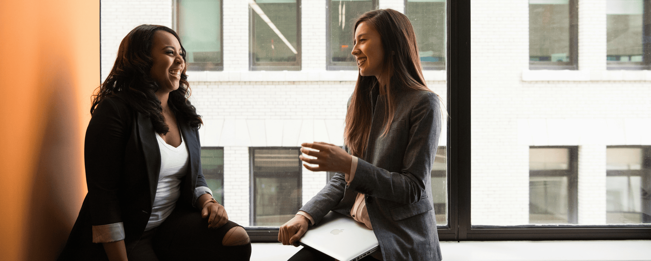 Two women are sitting by a large window, engaged in a friendly conversation in a professional setting, with one holding a laptop.