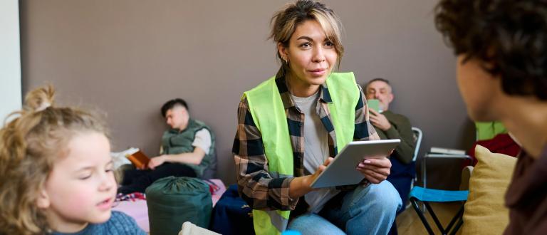 Une femme en gilet de sécurité tient une tablette et parle à des personnes dans un refuge, tandis que d'autres se reposent ou lisent en arrière-plan.