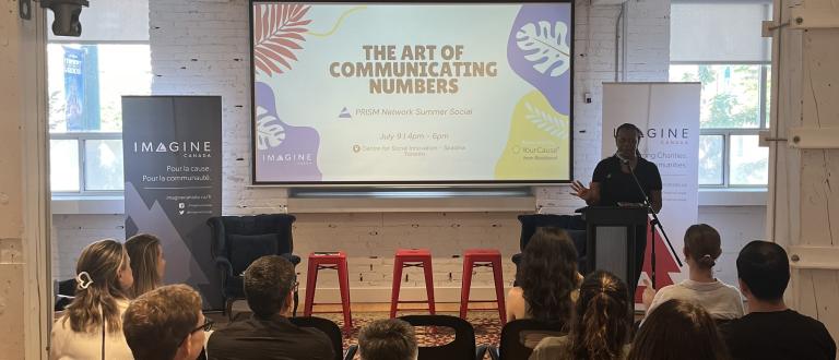 A speaker stands at a podium addressing an audience at an Imagine Canada event, with a screen behind them reading “The Art of Communicating Numbers” and event details for a PRISM Network Summer Social.