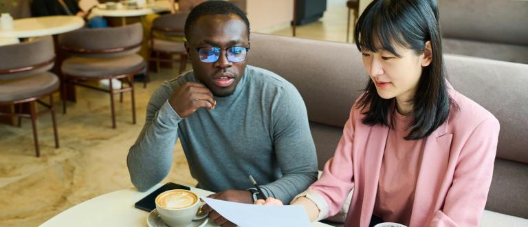 Two professionals meeting in a café, reviewing printed documents together over coffee.