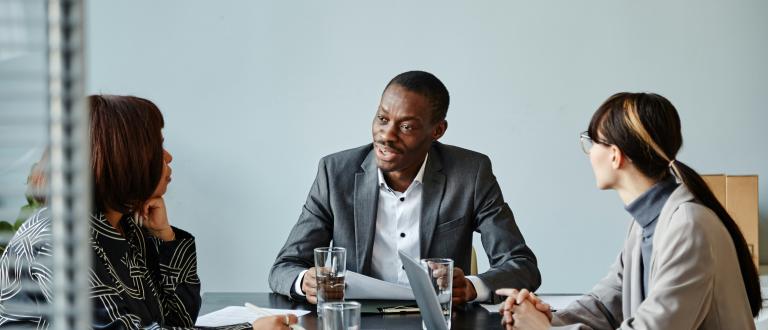 Three professionals sit at a table in a meeting room, engaged in discussion. One man in a gray suit speaks while two colleagues listen, taking notes and using laptops and documents.