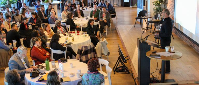 Bruce MacDonald parle à un podium devant un large public assis dans un espace moderne et lumineux, avec des tables rondes et des écrans de présentation.