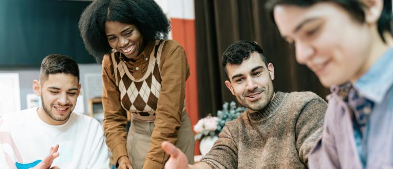 Un groupe de jeunes adultes assis autour d'une table pratiquent ensemble la poterie. L'un d'eux se lève et sourit tandis que les autres façonnent l'argile de leurs mains dans une ambiance détendue et conviviale d'atelier.
