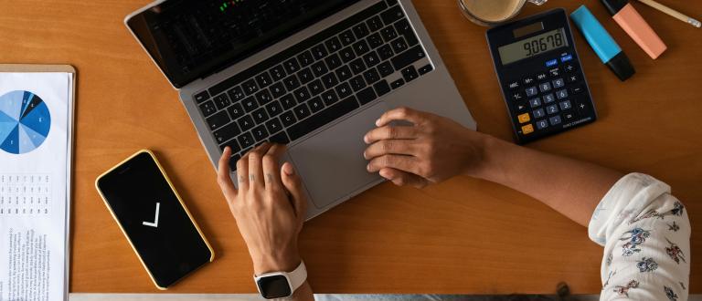 Overhead view of a person working on a laptop showing stock market charts, with a calculator, coffee, phone, and printed charts on a wooden desk.