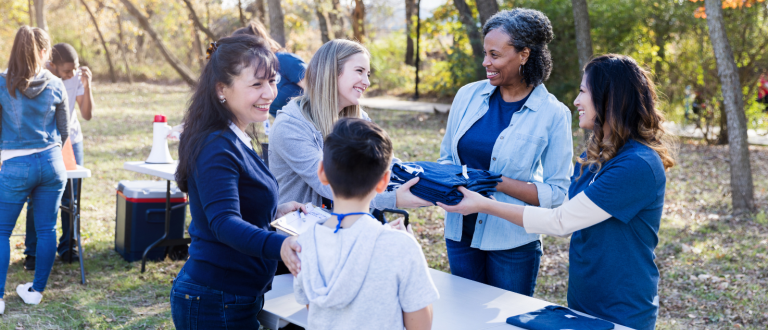 Un groupe de bénévoles souriants distribue des t-shirts bleus et des badges lors d'un événement communautaire en plein air.
