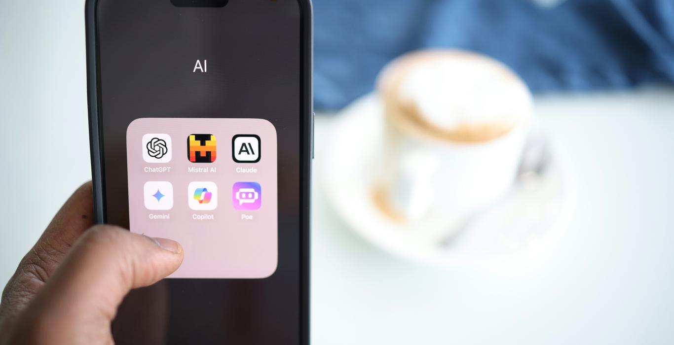 A person holds a smartphone displaying an AI folder with various app icons including ChatGPT, Midjourney, Claude, Gemini, CoPilot, and Poe.