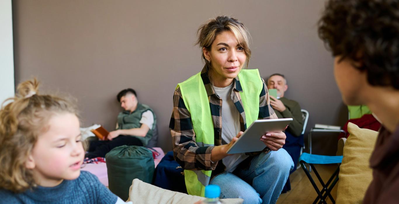 Une femme en gilet de sécurité tient une tablette et parle à des personnes dans un refuge, tandis que d'autres se reposent ou lisent en arrière-plan.