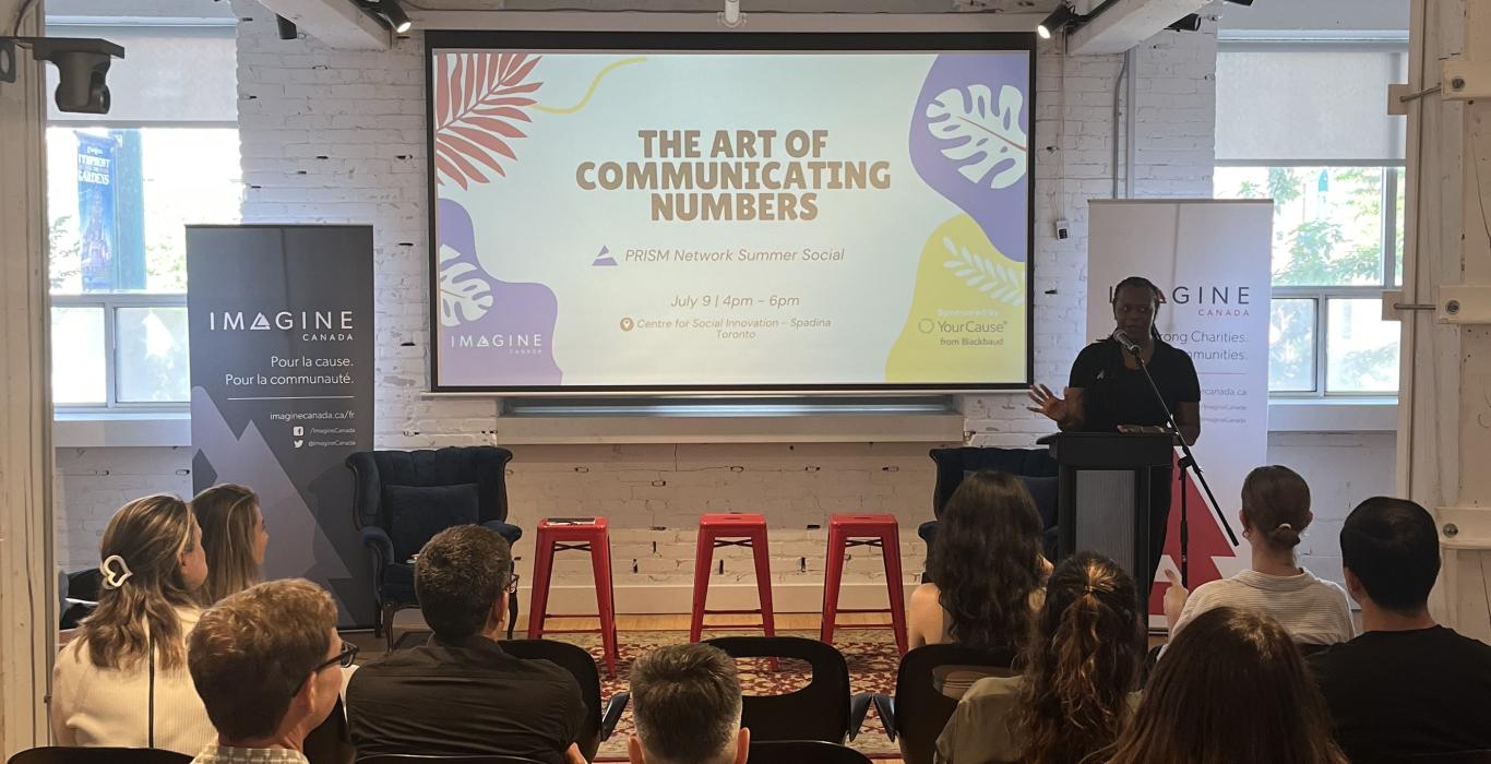 A speaker stands at a podium addressing an audience at an Imagine Canada event, with a screen behind them reading “The Art of Communicating Numbers” and event details for a PRISM Network Summer Social.