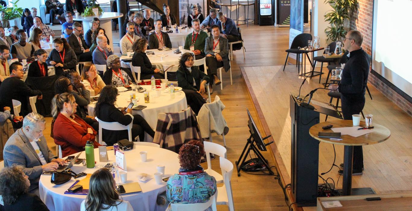 Bruce MacDonald parle à un podium devant un large public assis dans un espace moderne et lumineux, avec des tables rondes et des écrans de présentation.