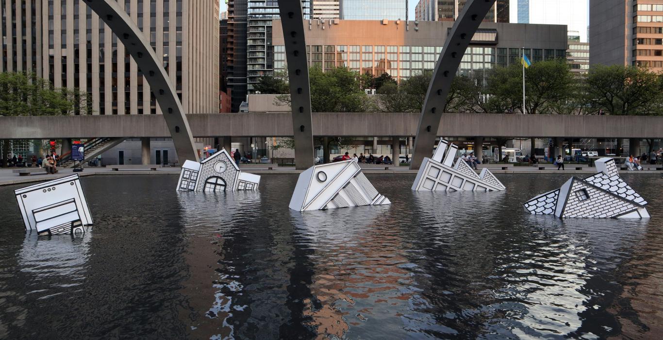 Sculptures de maisons submergées de style bande dessinée dans un bassin public, avec des immeubles et des arches en arrière-plan.