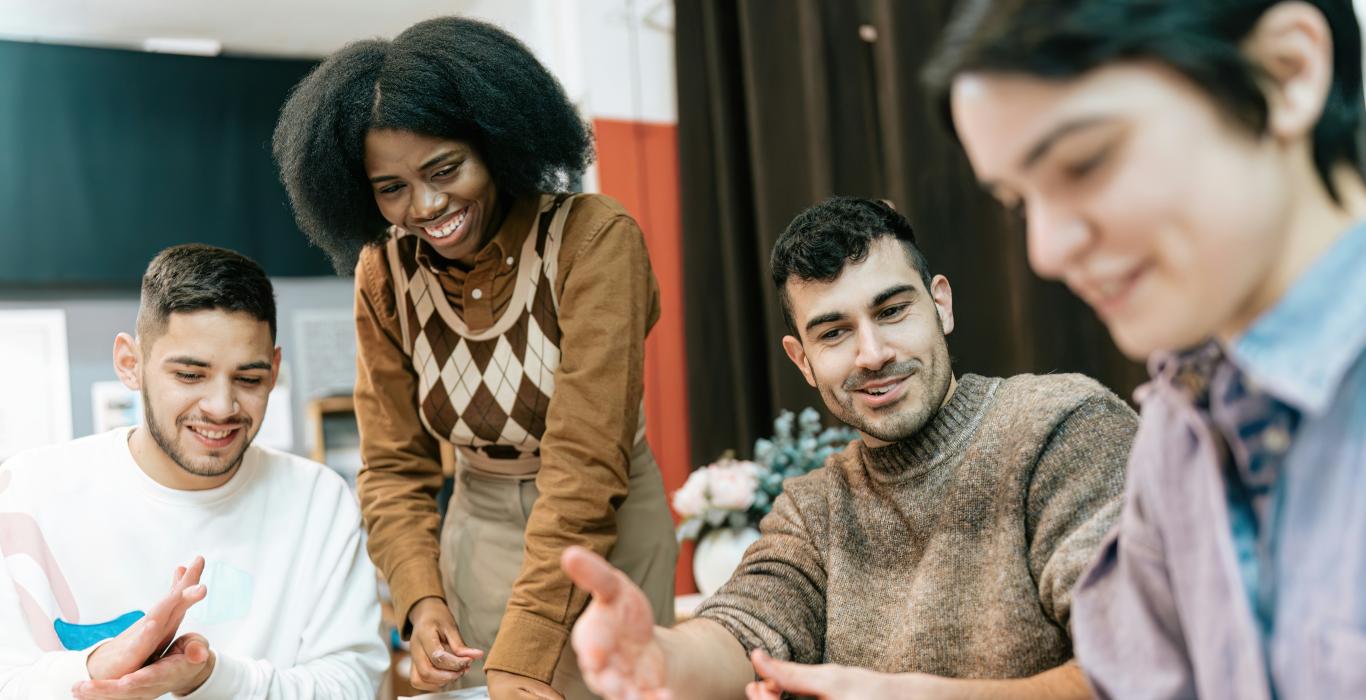 A group of young adults sitting around a table doing clay art together. One person stands and smiles while others shape clay with their hands in a relaxed, friendly workshop setting.