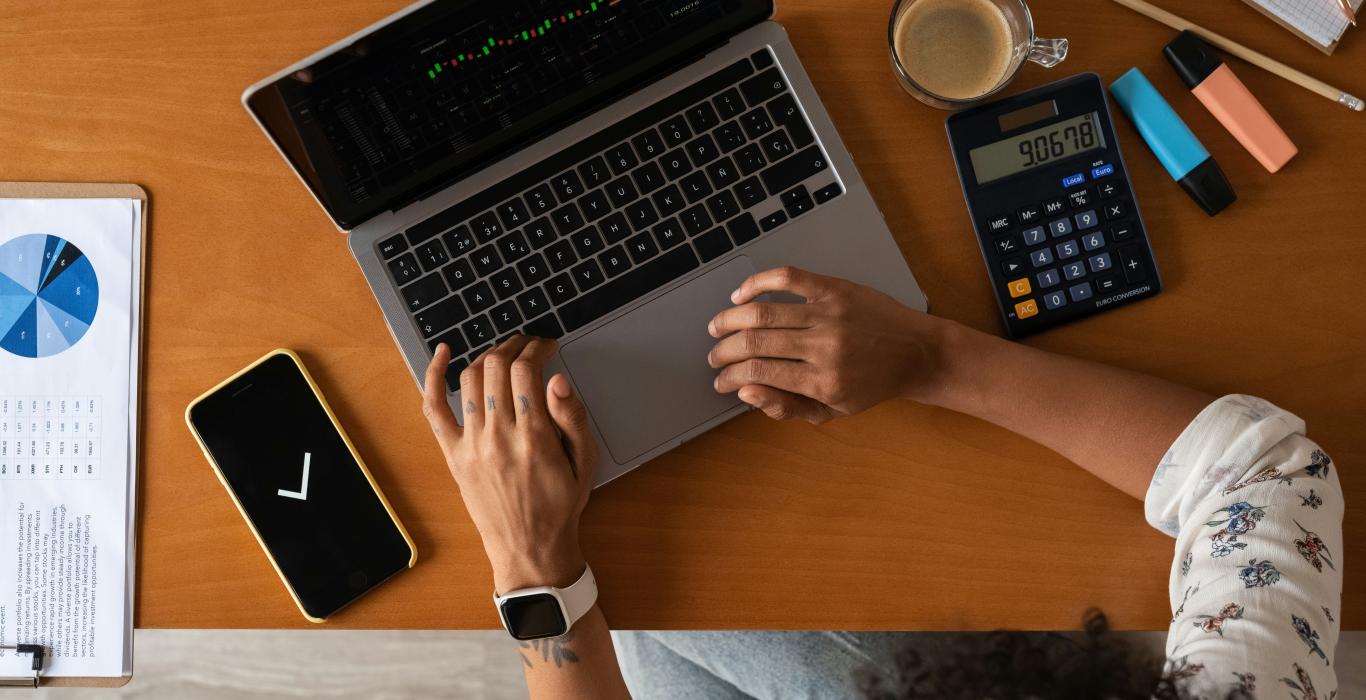 Overhead view of a person working on a laptop showing stock market charts, with a calculator, coffee, phone, and printed charts on a wooden desk.
