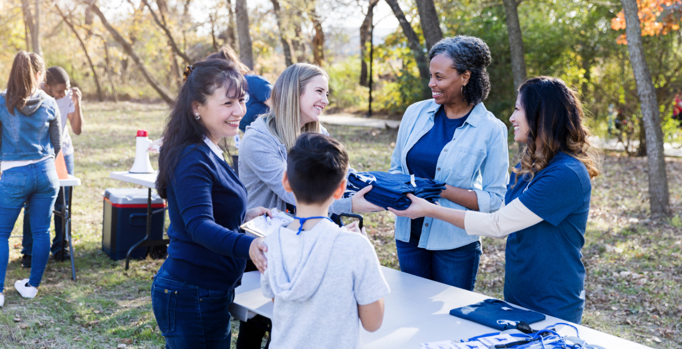 People gather outdoors at a picnic table in a park, smiling and handing out or receiving small items while others chat in the background among trees with autumn foliage.