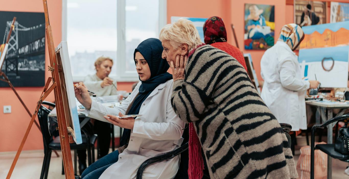 Une femme peint sur une toile, sous le regard d’une femme plus âgée, lors d’un atelier artistique en groupe.