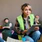 A woman wearing a safety vest holds a tablet while talking to people in a shelter setting; others rest or read in the background.
