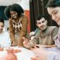 A group of young adults sitting around a table doing clay art together. One person stands and smiles while others shape clay with their hands in a relaxed, friendly workshop setting.